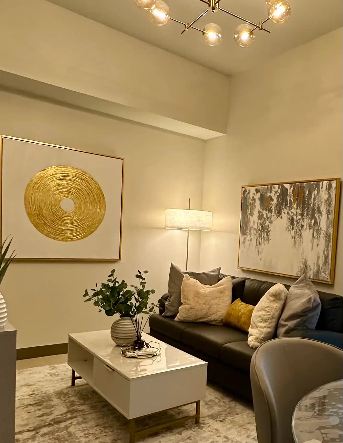 Luxury modern interior design installation featuring a large square metallic gold relief circular sunburst art piece and an abstract textured painting in gold, white, and charcoal grey hanging above a black leather living room sofa.