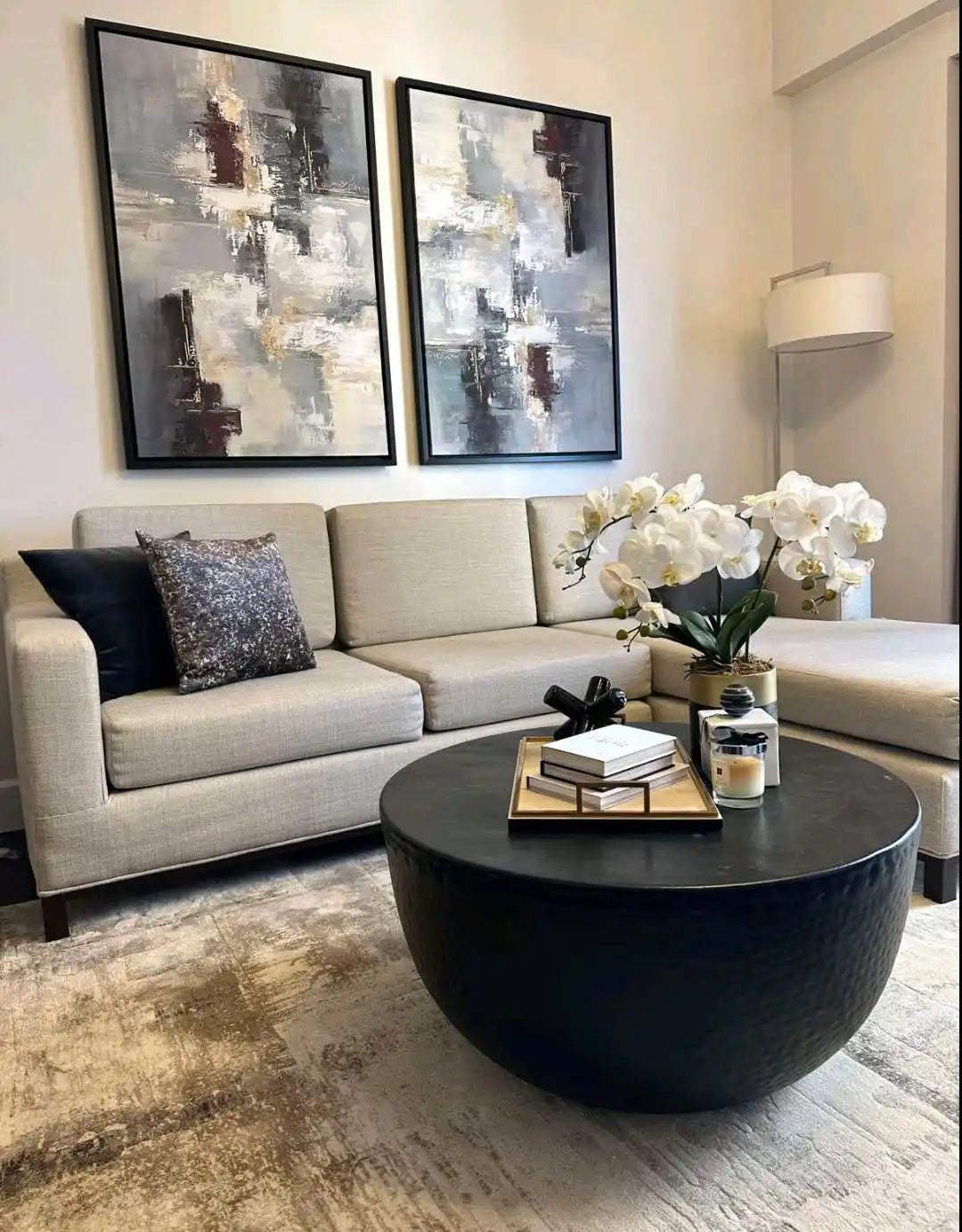 Modern minimalist abstract diptych canvas prints showcased in a real home installation, featuring heavy impasto textures and bold palette knife strokes in grey, charcoal black, white, and metallic gold hanging above a beige living room sofa.
