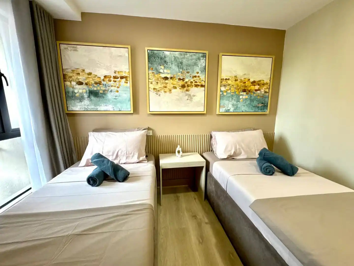 Modern minimalist abstract triptych canvas prints showcased in a real hotel installation, featuring coastal ocean horizon lines and abstract metallic splashes in teal blue, white, beige, and gold hanging above twin beds.