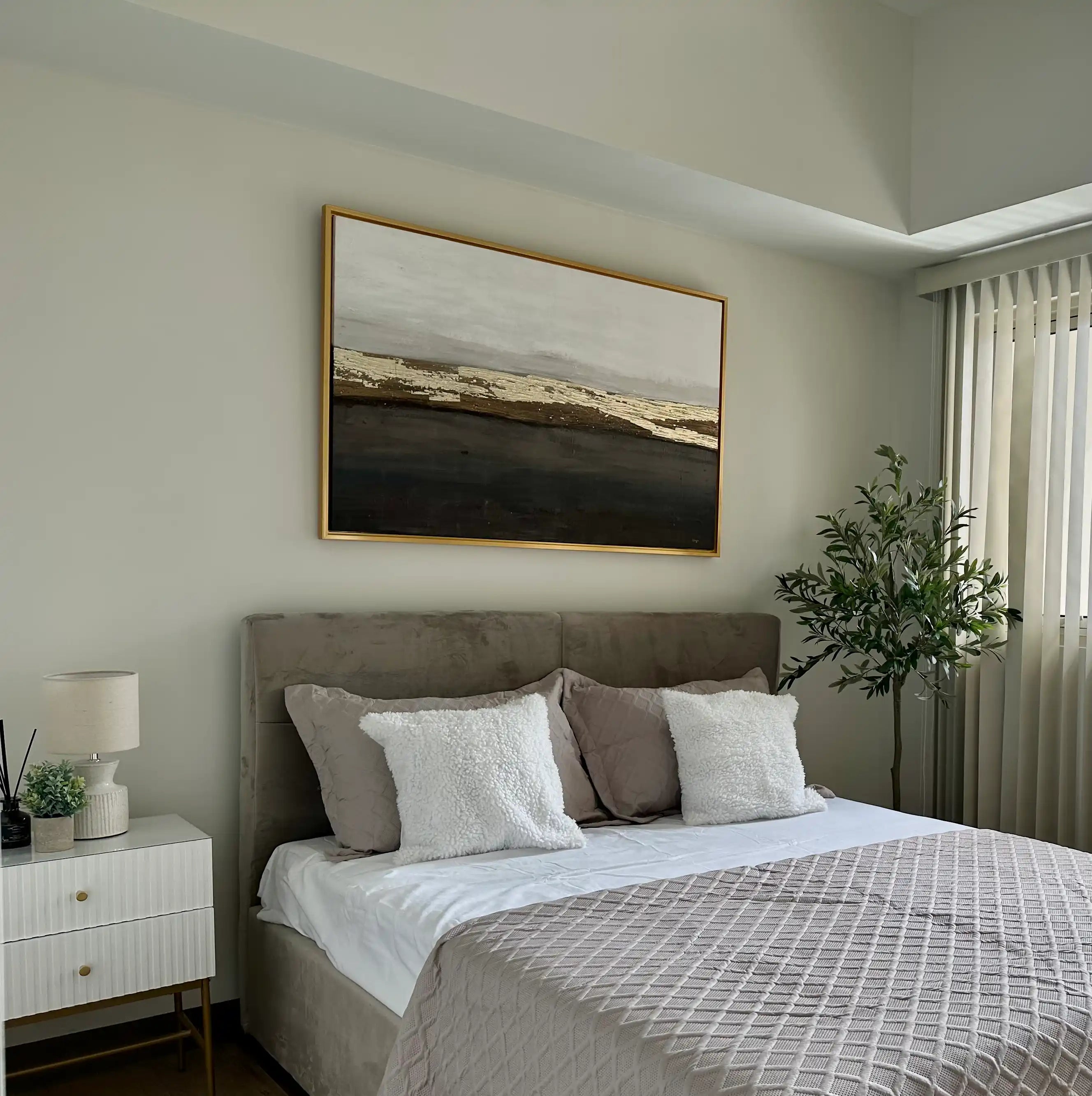 Oversized minimalist abstract landscape canvas print showcased in a real bedroom installation, featuring layered textured horizon blocks in black, charcoal grey, beige, and metallic gold foil hanging above a modern tufted headboard.